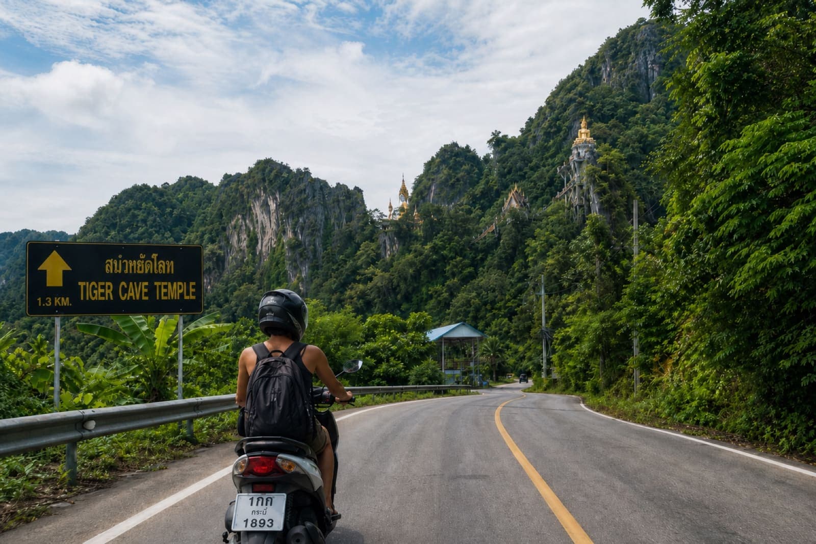 Helmeted scooter ride on a road toward Tiger Cave Temple area in Krabi with lush green hills