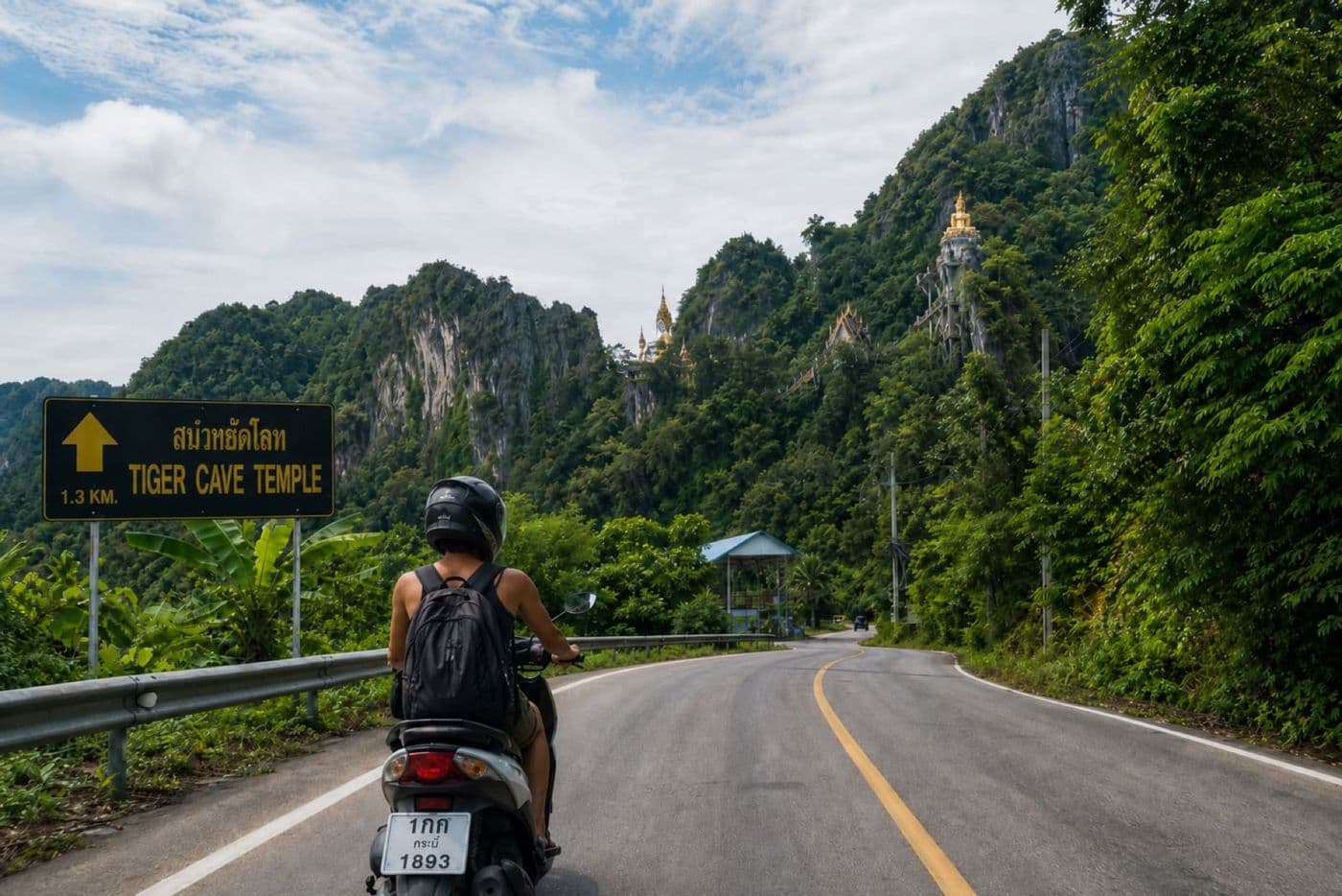Helmeted scooter ride on a road toward Tiger Cave Temple area in Krabi with lush green hills